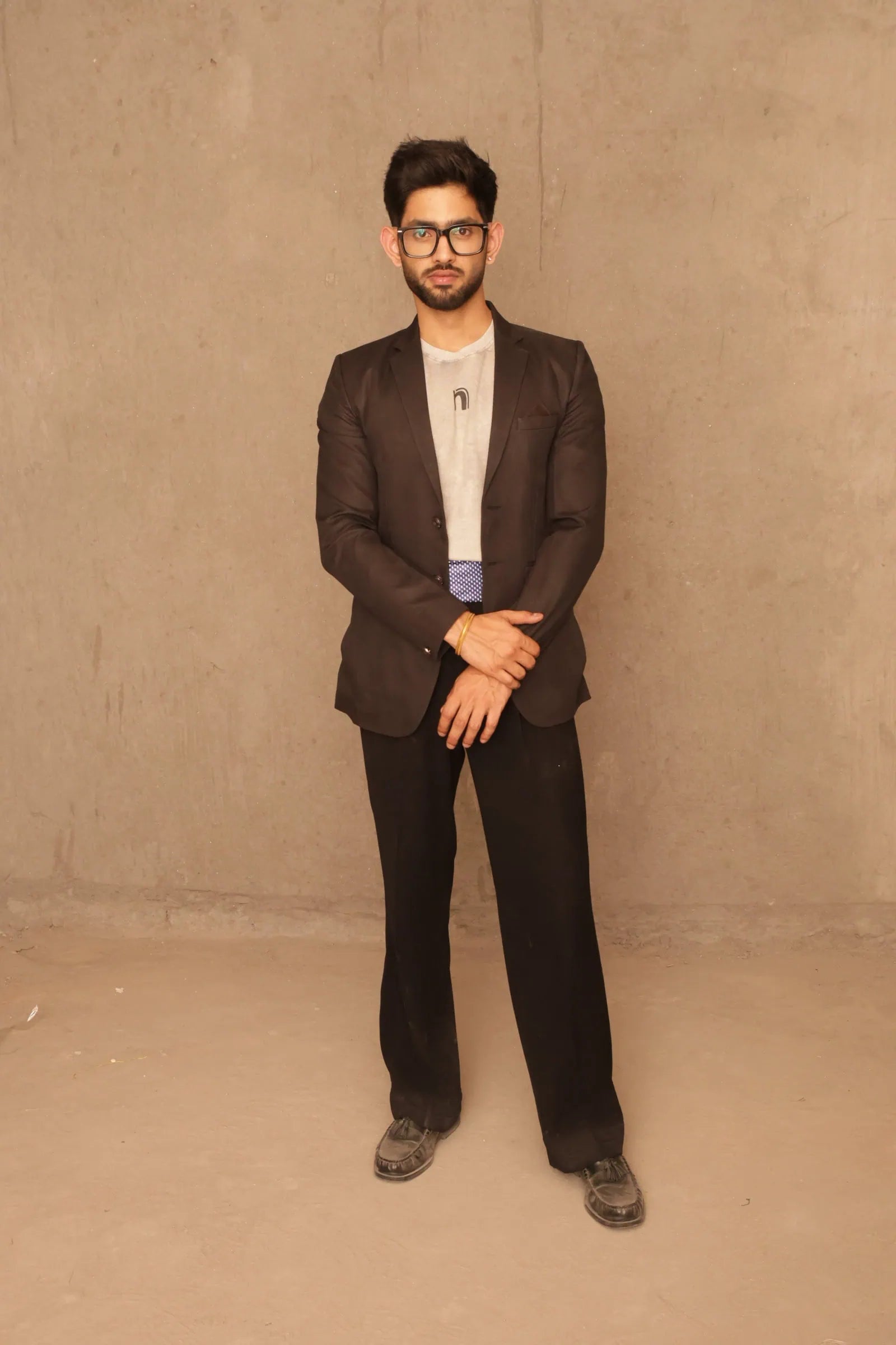 Man wearing a brown blazer over a light shirt with dark pants against a beige wall.