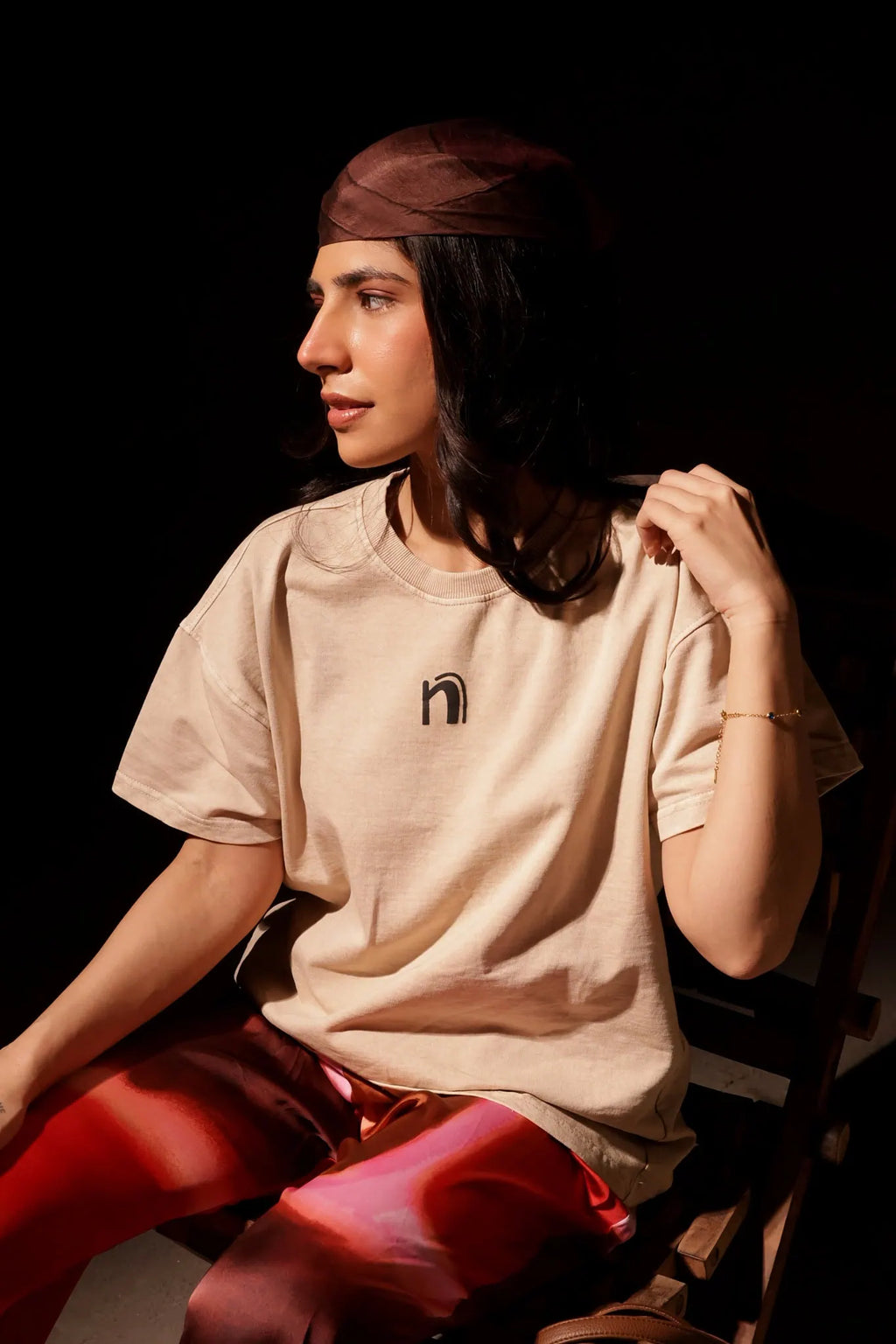 Person wearing a beige t-shirt with a logo and red pants sitting against a dark background