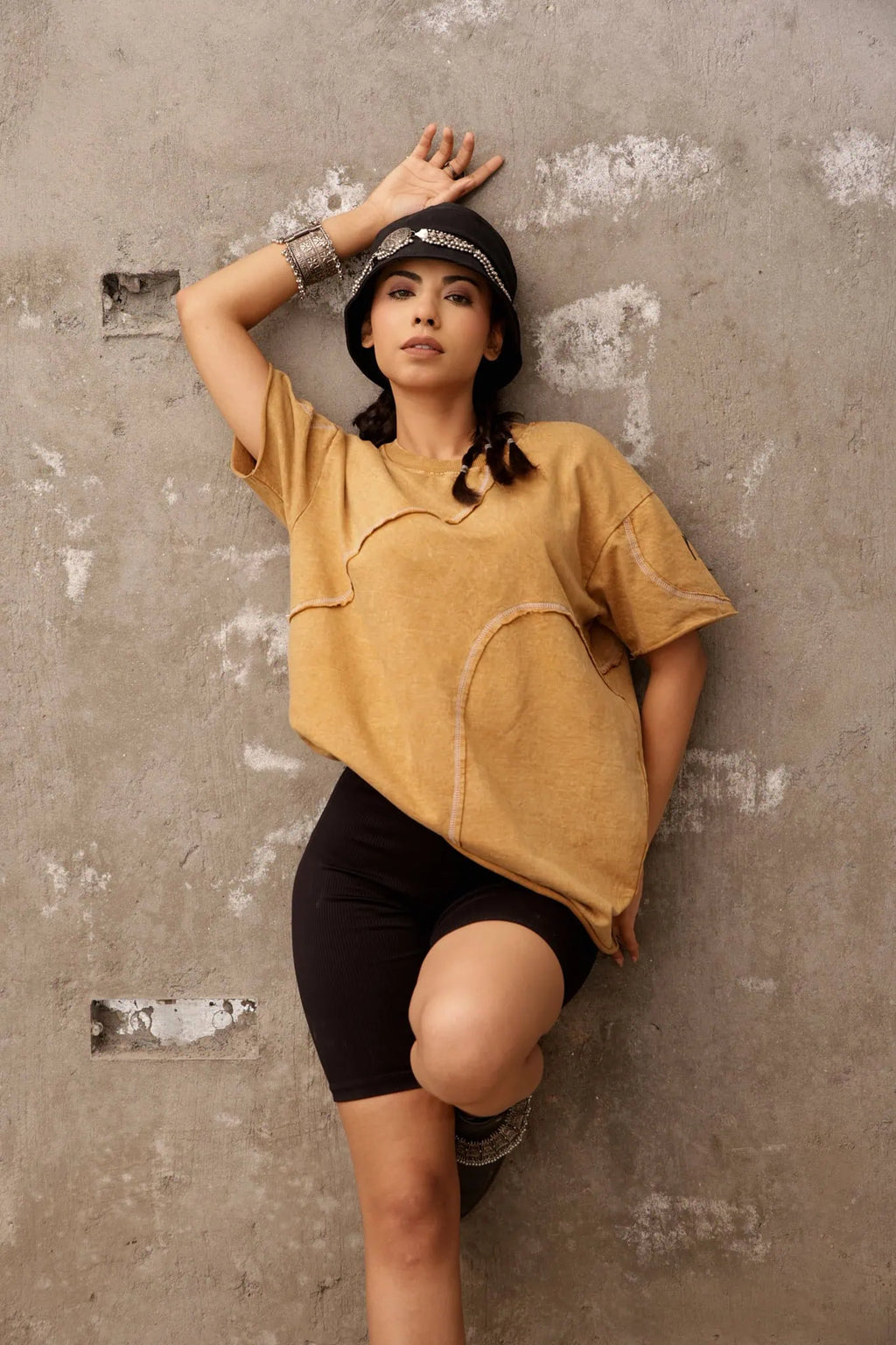 Woman in mustard yellow oversized shirt and black shorts leaning against a textured wall.