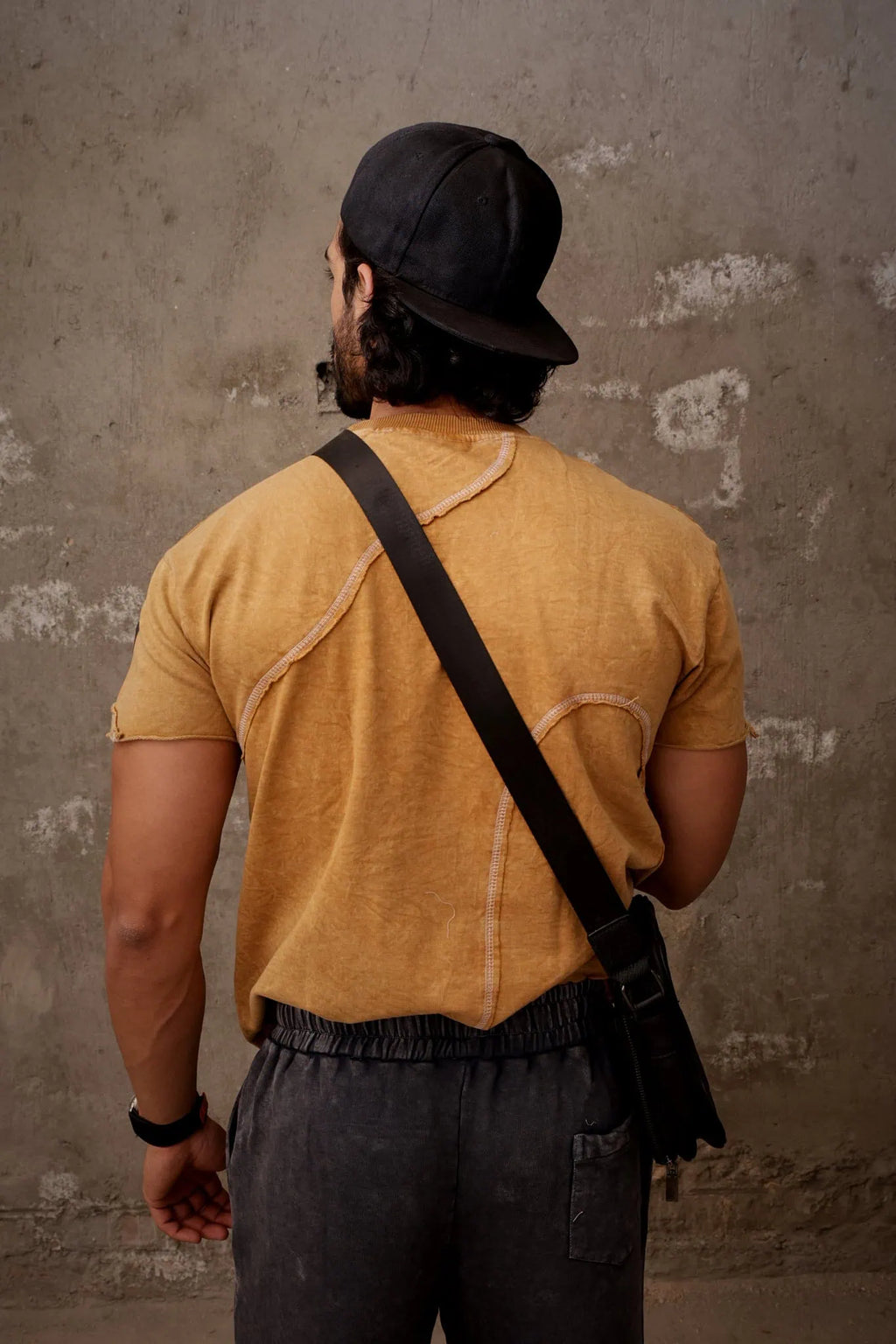 Man wearing a mustard yellow shirt and black cap, standing against a textured wall.