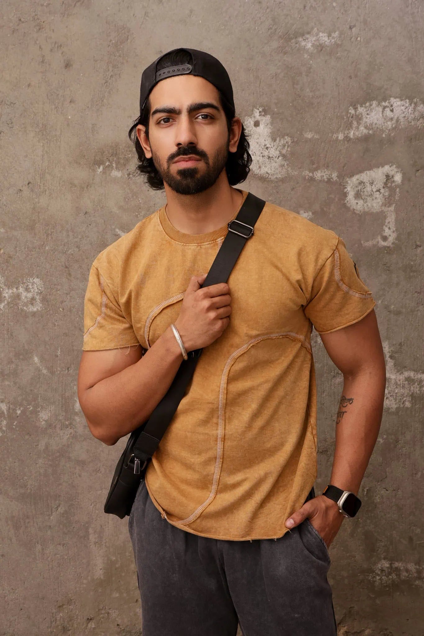 Man wearing a mustard yellow shirt and black cap against a textured wall.