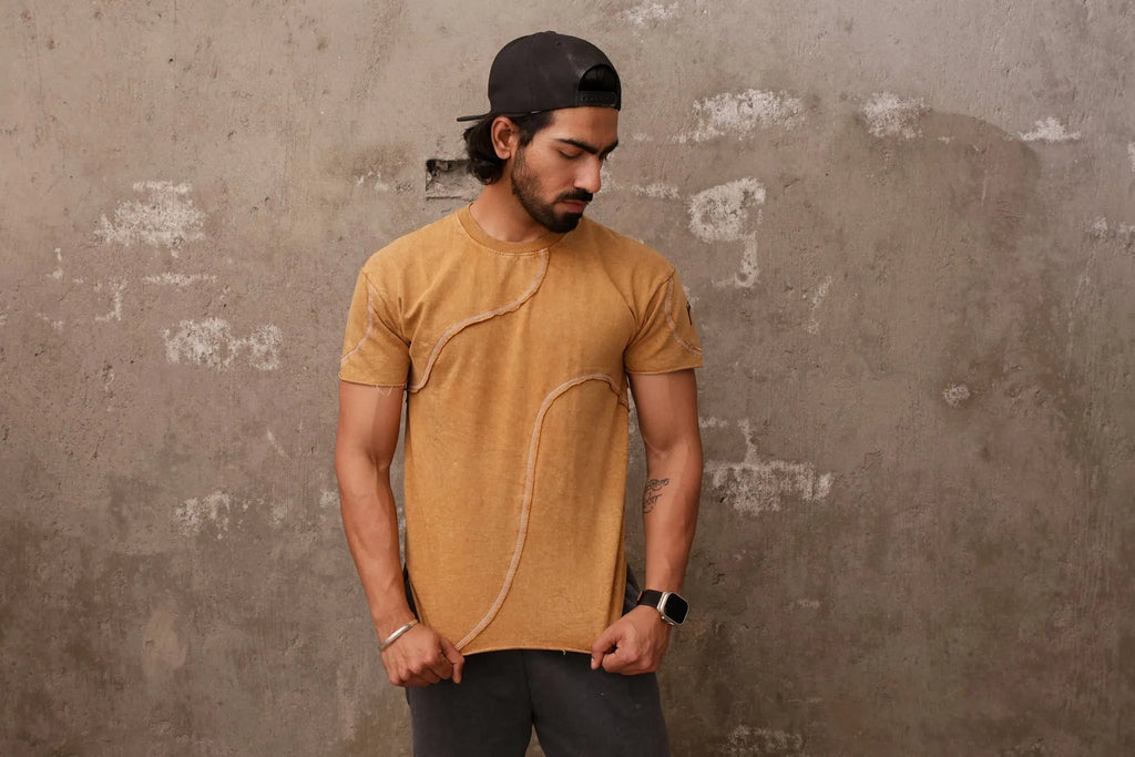 Man wearing a mustard yellow t-shirt and black cap against a textured wall.