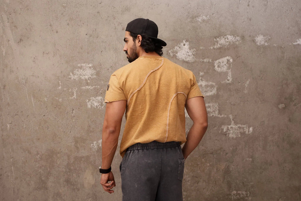 Man wearing a mustard yellow t-shirt and dark pants against a textured wall.