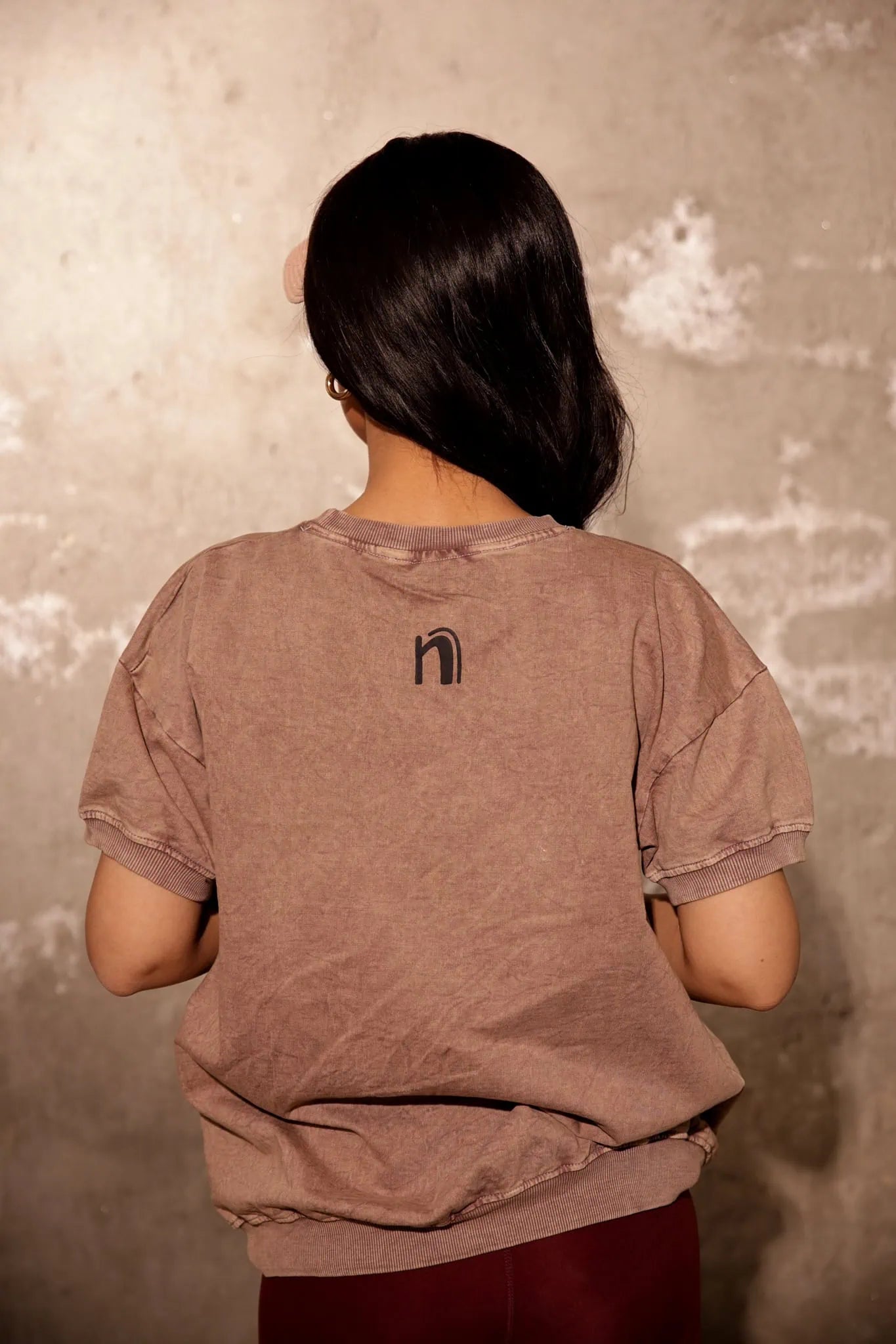 Person wearing a brown t-shirt with a logo on the back against a textured wall.