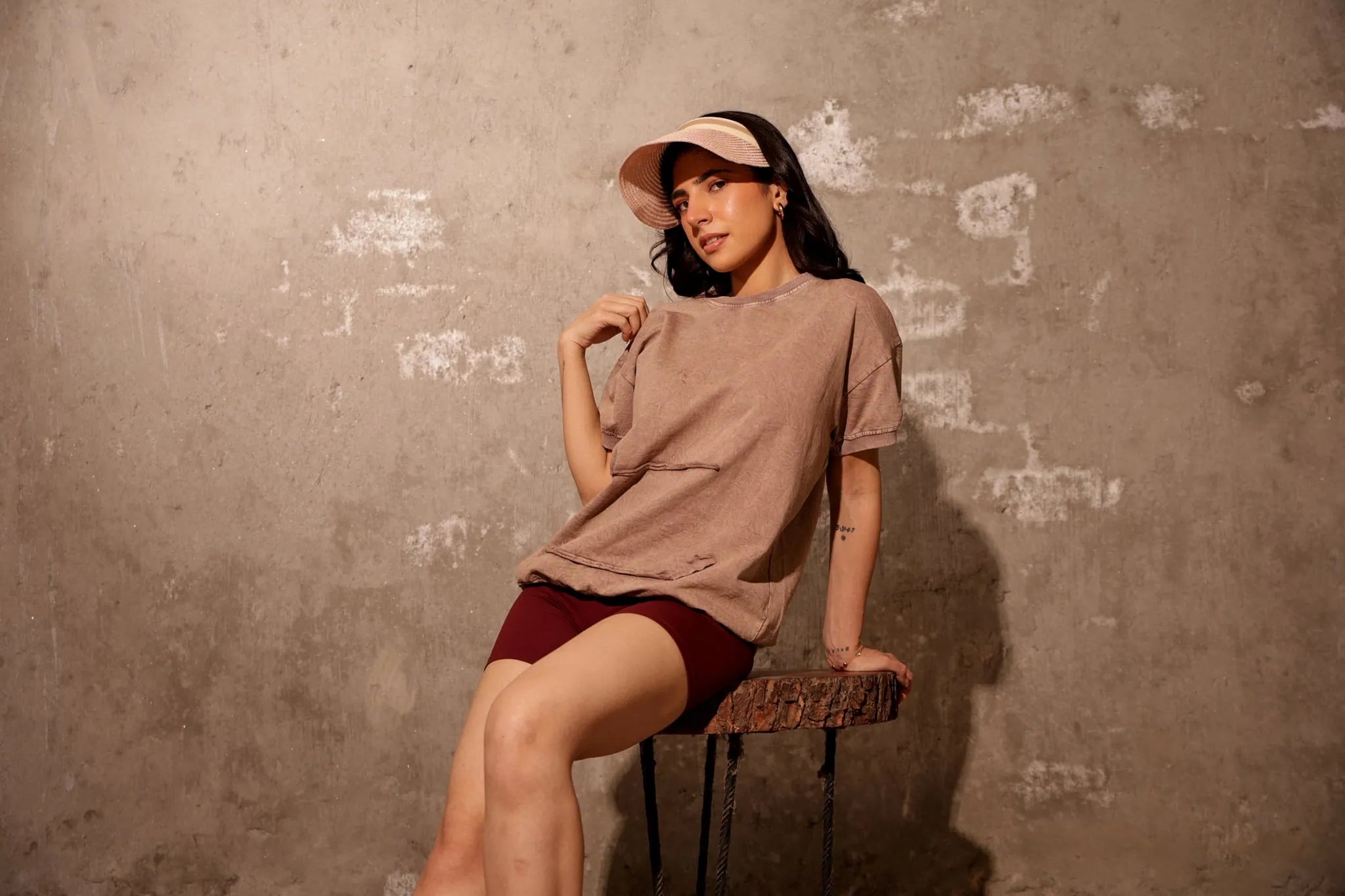 Woman sitting on a chair wearing a beige t-shirt and maroon shorts against a textured wall.