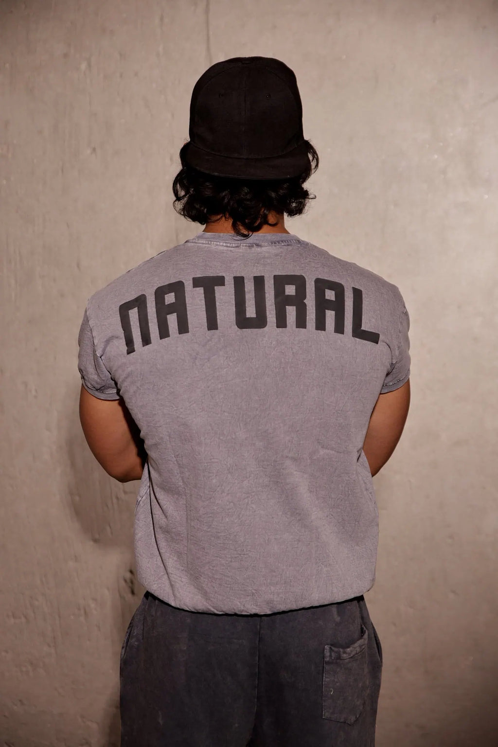 Person wearing a gray t-shirt with 'NATURAL' printed on the back against a plain wall.