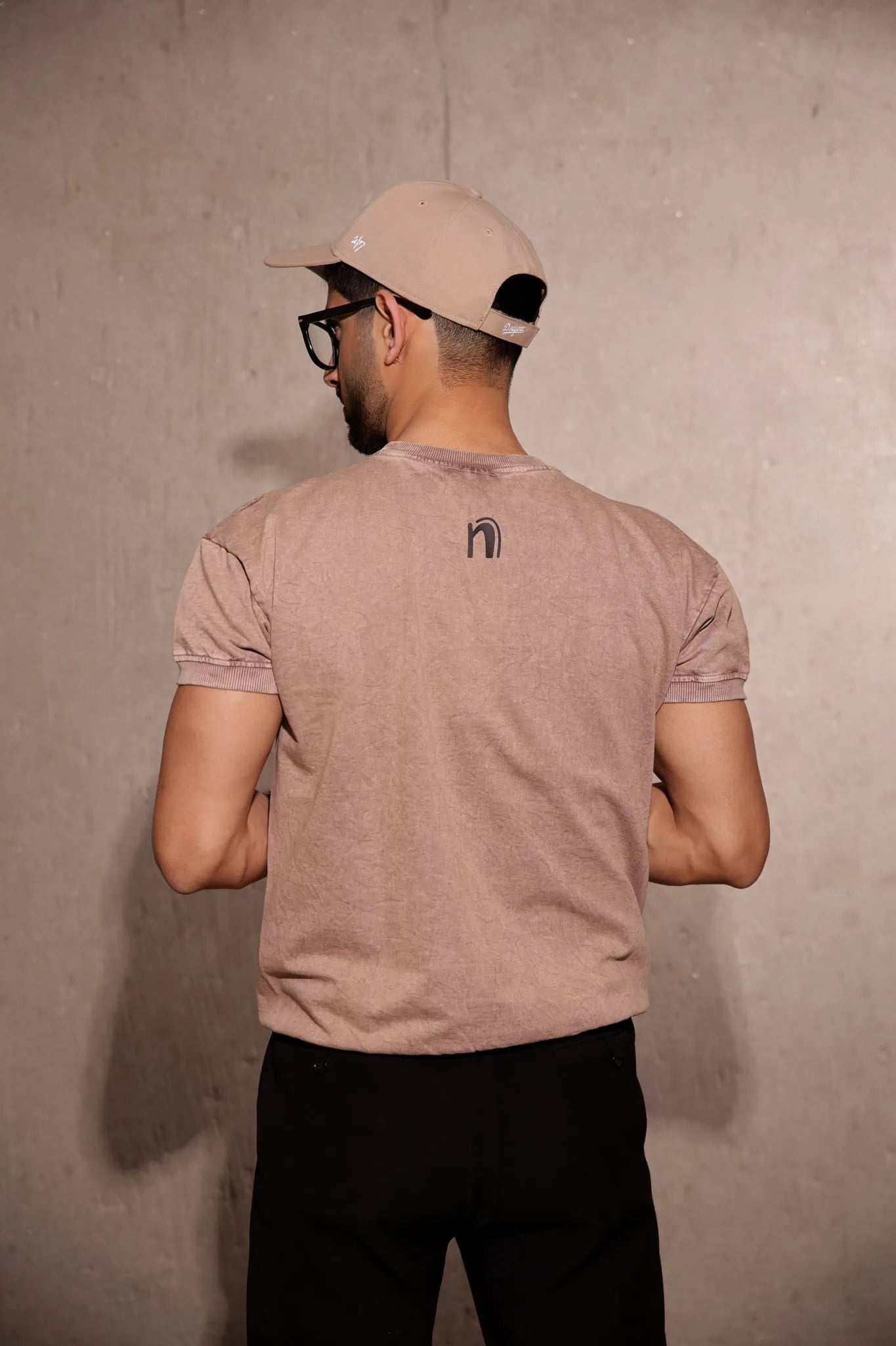 Man wearing a beige t-shirt with a logo, standing against a plain wall.