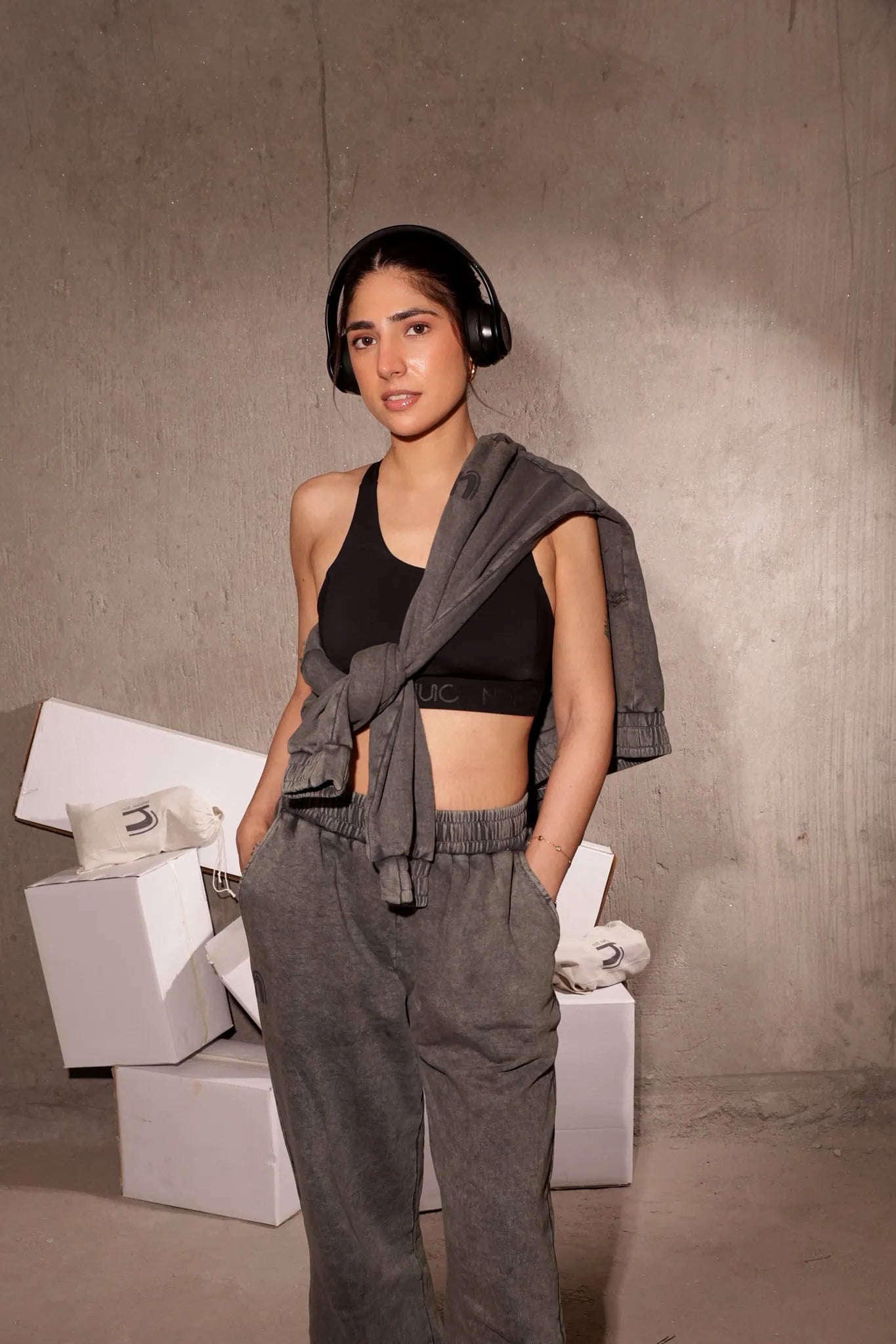 Woman in black sports bra and gray sweatpants standing against a concrete wall.