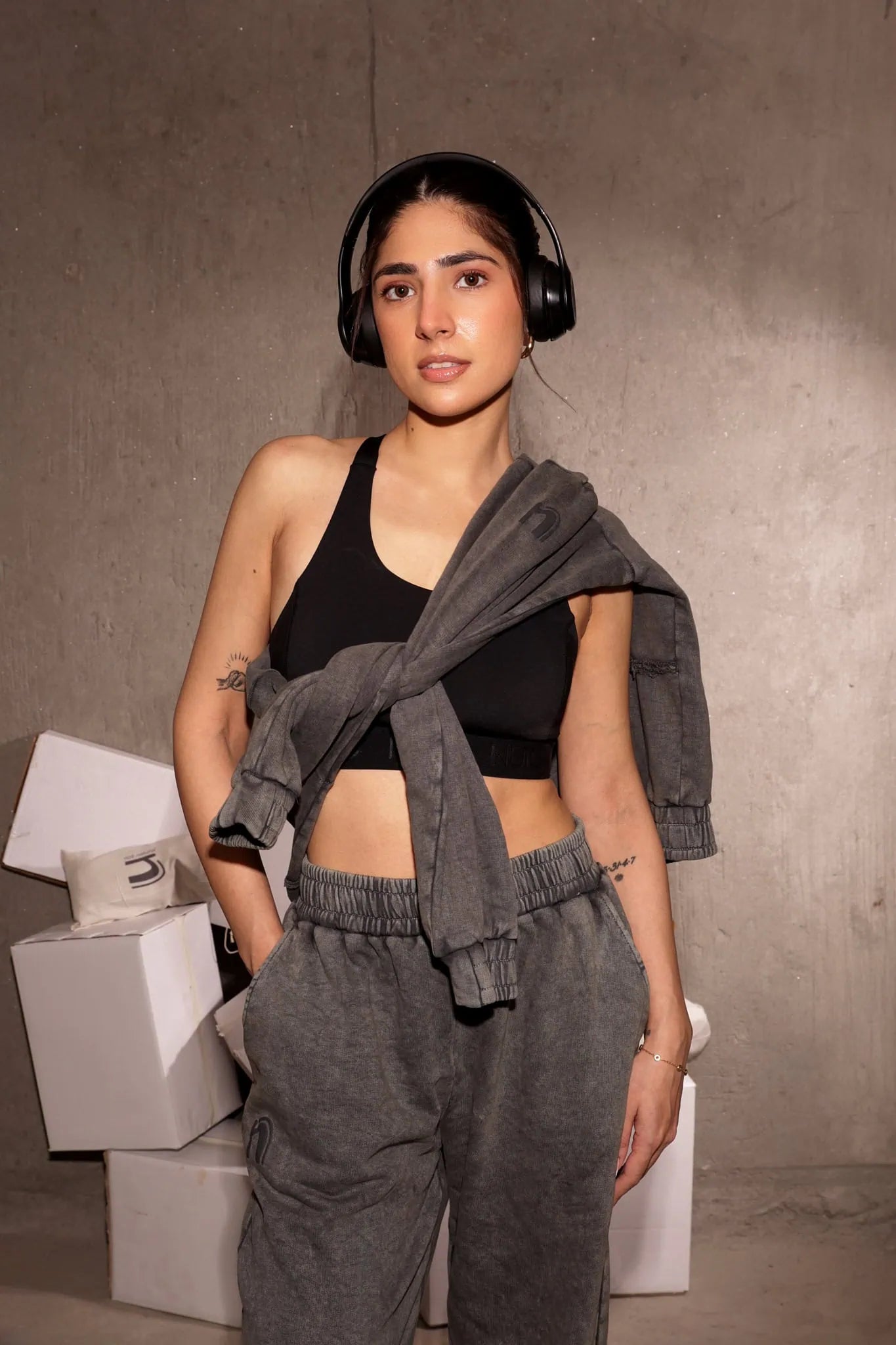 Woman wearing a black sports bra and gray sweatpants with headphones, standing against a neutral background.