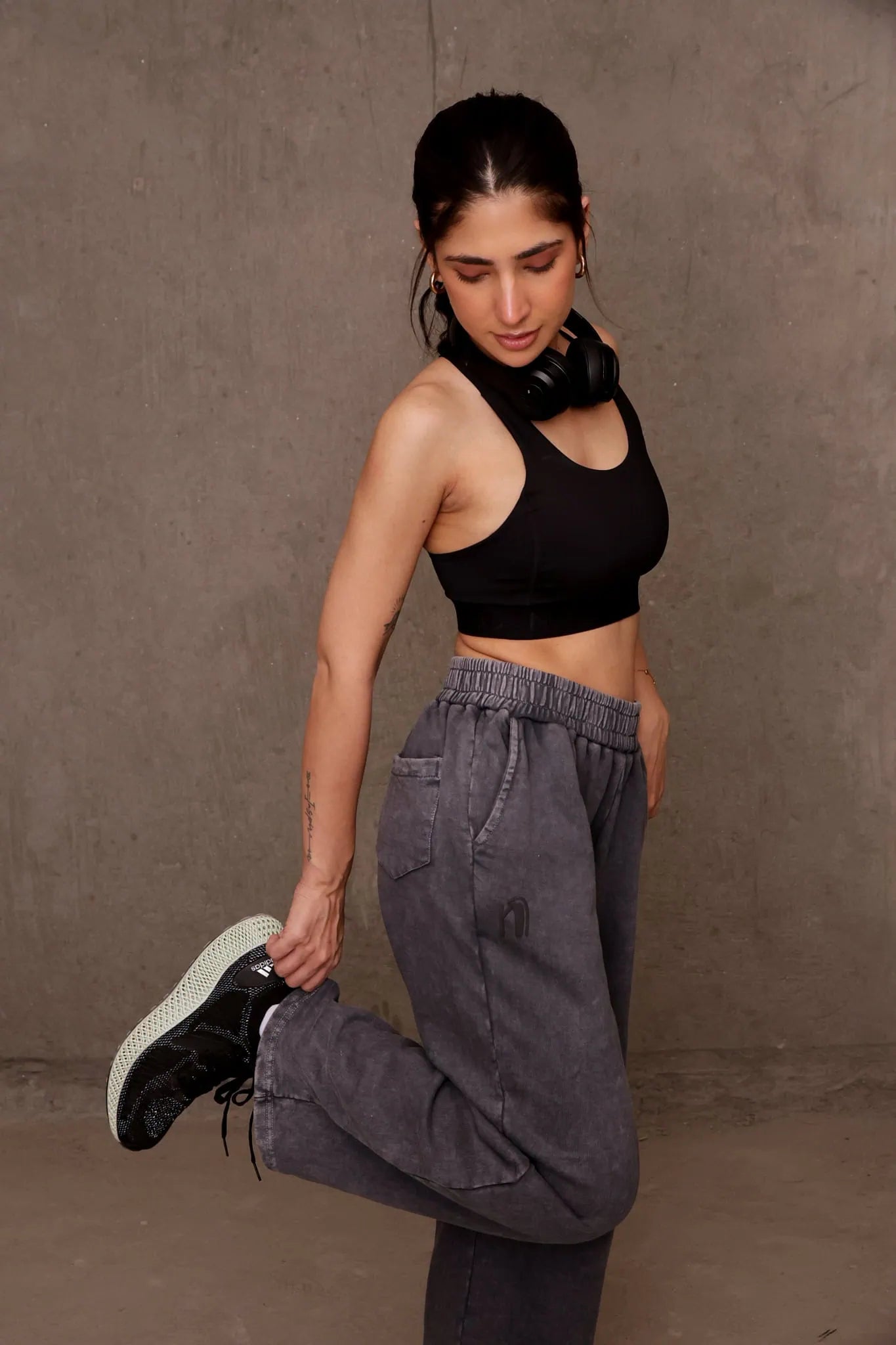 Woman in black sports bra and gray sweatpants posing against a brown wall.